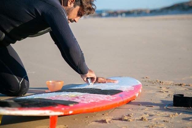 Fokussierter hübscher Surfer im Neoprenanzug-Polier-Surfbrett mit Wachs auf Sand