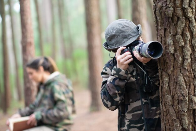 Fokussierter Junge mit Kamera im Wald. Schüler im Mantel und Panama beim Fotografieren. Undeutliche Mutter im Hintergrund. Kindheit, Natur, Freizeitkonzept
