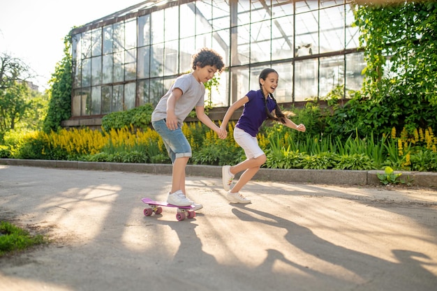 Fokussierter lockiger Junge, der lernt, das Skateboard zu fahren, begleitet von einem fröhlichen süßen Mädchen