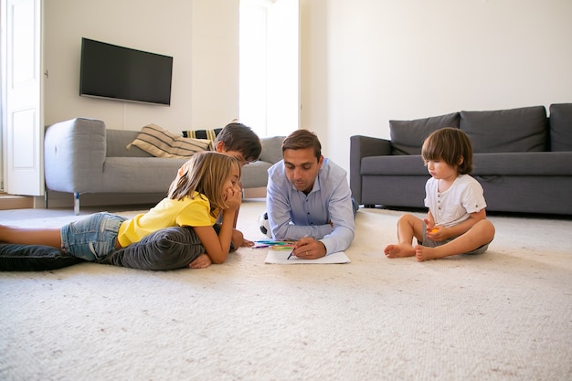 Fokussierter Vater und Kinder, die auf Teppich liegen und auf Papier malen. Liebender kaukasischer Vater, der mit Markern zeichnet und mit niedlichen Kindern zu Hause spielt. Konzept für Kindheit, Spielaktivität und Vaterschaft