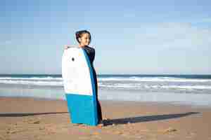 Kostenloses Foto fokussiertes kleines mädchen mit surfbrett am strand. dunkelhaariges kind in surfuniform an einem sonnigen tag, seitlich schauend. sport, freizeit, aktives lifestyle-konzept