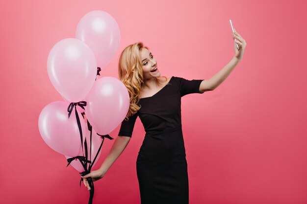 Formschön faszinierende junge Frau, die neues Telefon für Selfie verwendet. Lachendes Geburtstagskind, das Foto von sich selbst macht, während es großen Haufen Partyballons hält.