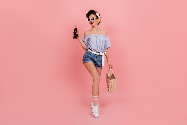 Formschönes Pinup-Mädchen, das eine Flasche Soda hält und lächelt. Atemberaubende europäische Frau mit Sommertasche, die auf rosa Hintergrund aufwirft.