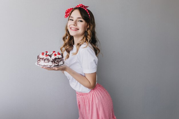 Formschönes weibliches Modell mit rotem Band, das süßen Kuchen hält. Innenaufnahme der blithesome lockigen Frau, die Geburtstagstorte hält.