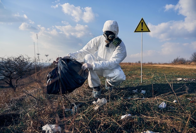Forscher im Schutzanzug, der an sonnigen Tagen im Freien Plastikmüll in schwarzen Abfallsack sammelt