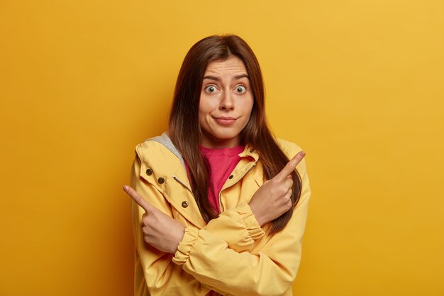 Foto der ahnungslosen zögernden Frau mit den dunklen Haarspitzen seitwärts, wählt zwischen zwei Optionen, hat Gesichtsausdruck überrascht, trägt Jacke, posiert gegen gelbe Wand, sagt besser, um zu schauen