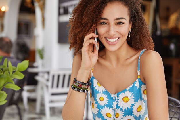 Foto der angenehm aussehenden afroamerikanischen Frau mit fröhlichem Ausdruck, ruft Freund über Handy an, genießt Freizeit während des Sommertages