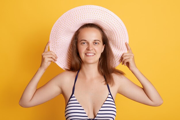Foto der angenehm aussehenden jungen Frau im gestreiften Bikini und im Strohhut, schaut mit glücklichem Ausdruck