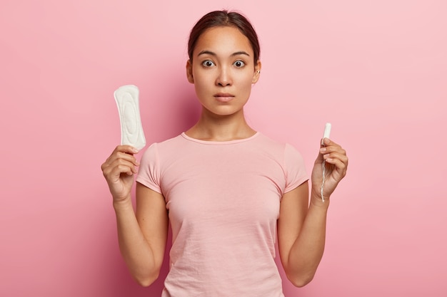 Foto der attraktiven asiatischen Frau hält Baumwolltampon und Damenbinde, hat überraschenden Blick, monatlichen Menstruationszyklus, trägt rosa T-Shirt. Schutz für die Gesundheit von Frauen. Düngungskonzept