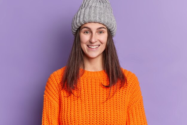 Foto der attraktiven europäischen Frau mit dem langen dunklen Haar angenehmes Lächeln, das glücklich ist, Winterferien zu haben, trägt orange gestrickten Pullover und Hut.