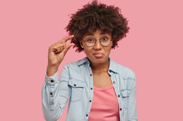 Foto der attraktiven jungen Frau mit Afro-Haarschnitt zeigt etwas sehr kleines oder winziges, Gesten mit der Hand, hat dunkle Haut, gekleidet in Jeansjacke, isoliert über rosa Wand. Es ist zu klein