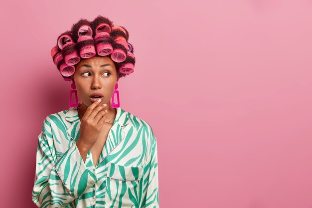 Foto der dunkelhäutigen Frau bekommt Haare gekräuselt, trägt Lockenwickler und macht Frisur zu Hause, hält die Hand auf offenem Mund, trägt Freizeitkleidung, posiert gegen rosa Wand, leere Leerstelle beiseite