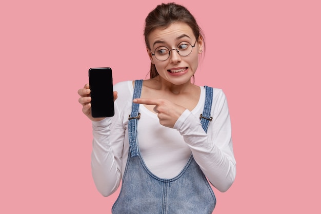 Foto der emotionalen jungen Frau zeigt auf Bildschirm des modernen Gadgets, zögert, ob man Smartphone kauft, sieht verwirrt aus