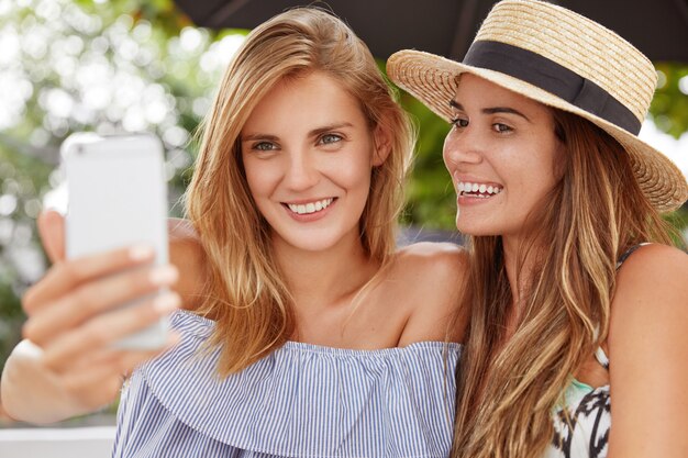 Foto der entzückenden jungen Frau mit hellem Haar verbringt Freizeit in Gesellschaft ihrer besten Freundin, hält Smartphone für Selfie, posiert zusammen in der Cafeteria im Freien, hat positive Ausdrücke