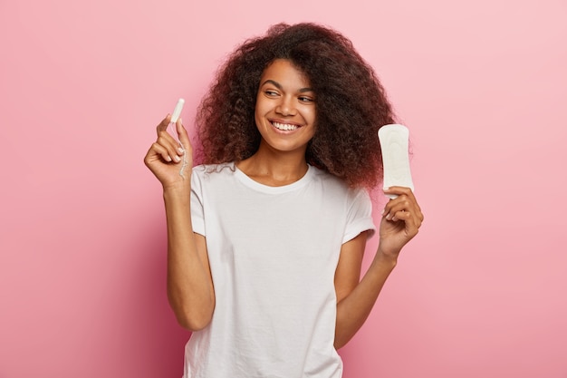 Foto der entzückten afroamerikanischen Frau hält Tampon und Damenbinde, gekleidet in weißem T-Shirt, lokalisiert über rosa Wand. Frauen, pms