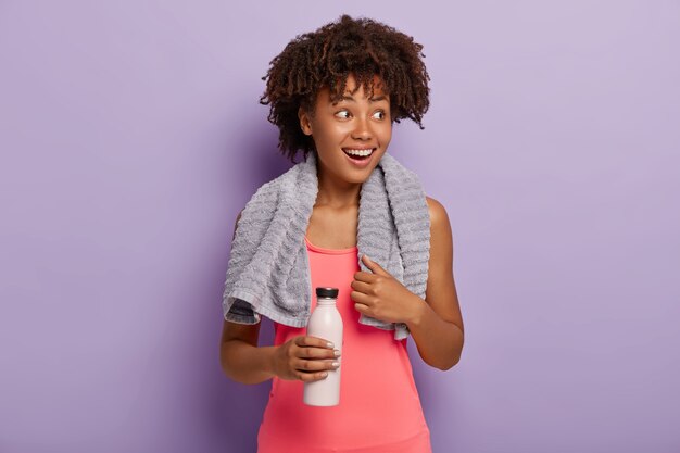 Foto der entzückten dunkelhäutigen Sportlerin hat Afro-Haarschnitt, schaut mit einem Lächeln zur Seite, trägt ein rosa Oberteil, trägt eine Flasche, trinkt Wasser, während sie sich während des Trainings durstig fühlt, trainiert drinnen.