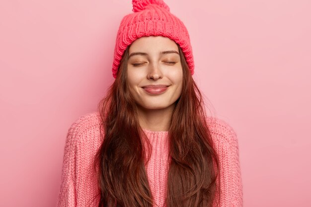 Foto der erfreuten langhaarigen erfreuten kaukasischen Frau hält die Augen geschlossen, lächelt sanft, hat gesunde Haut, trägt warme Strickmütze und Pullover, posiert vor rosa Hintergrund, stellt sich etwas Schönes vor