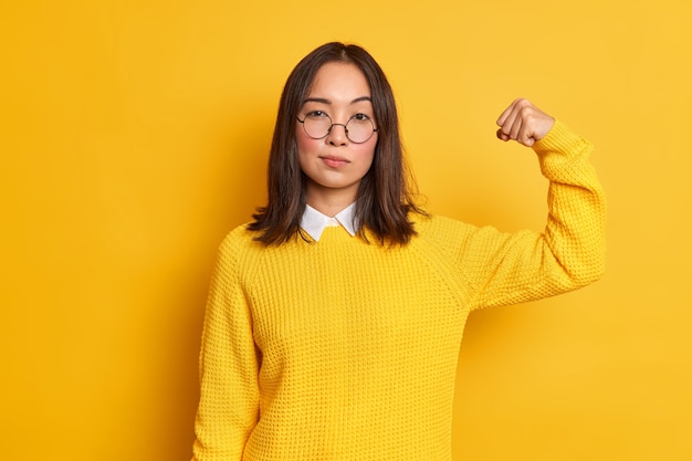 Foto der ernsthaften brünetten asiatischen Frau hebt den Arm und zeigt, dass ihre Stärke starke Muskeln hat, steht selbstbewusst drinnen, trägt gelben Pullover und runde optische Brille. Frauenpower-Konzept