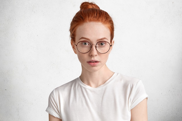 Foto der ernsthaften klugen rothaarigen Studentin mit Ingwerhaarknoten, trägt runde Brille, kommt auf Vorlesung an der Universität, isoliert über Weiß