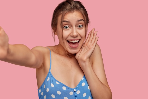 Foto der erstaunten fröhlichen jungen Frau mit glücklichem überglücklichem Ausdruck, hält Hand nahe Wange, gekleidet in gepunktetes Kleid, posiert gegen rosa Wand. Inhalt emotionales europäisches Mädchen posiert innen
