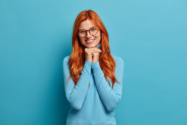 Foto der europäischen Frau mit roten Haaren hält Hände unter Kinn drückt positive Emotionen aus trägt optische Brille und lässigen Rollkragenpullover.