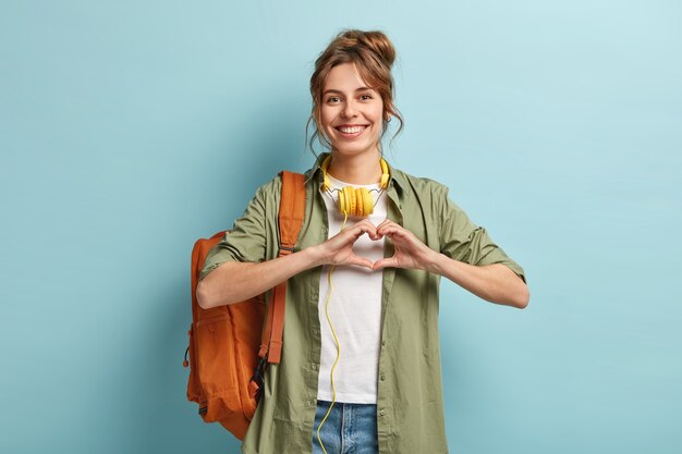 Foto der freundlichen hübschen weiblichen Reisenden macht Herzgeste über Brust, drückt Liebe zu Menschen aus, reist nur mit einem Rucksack