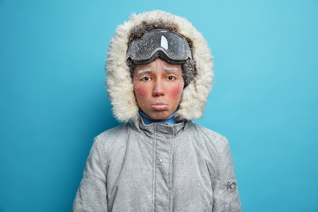 Foto der frustrierten gefrorenen Frau hat rdd Gesicht bedeckt mit Raureif verbringt Zeit im Freien während des frostigen Wintertages trägt Snowboardbrille und warme Thermojacke.