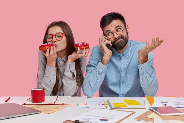 Foto der glücklichen Frau genießt Kaffeepause, isst leckeren Donut, studiert zusammen mit Gruppenmitglied, das über Handy spricht, hat ahnungslosen Ausdruck