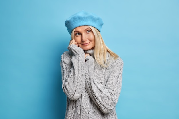 Foto der gut aussehenden Frau mittleren Alters hat verträumten nachdenklichen Ausdruck trägt gemütlichen wintergrauen Pullover und blaue Baskenmütze fühlt sich optimistisch in guten Erwartungen, die während des regnerischen Herbsttages ausgehen werden