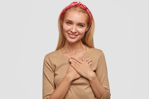 Foto der hübschen blonden lächelnden jungen Frau mit ansprechendem Blick, hält Hände auf Brust, drückt gutes Gefühl aus