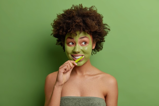 Foto der jungen Afroamerikanerin wendet grüne Gesichtsmaskenpinsel an, die Zähne verwenden, verwendet Zahnbürste