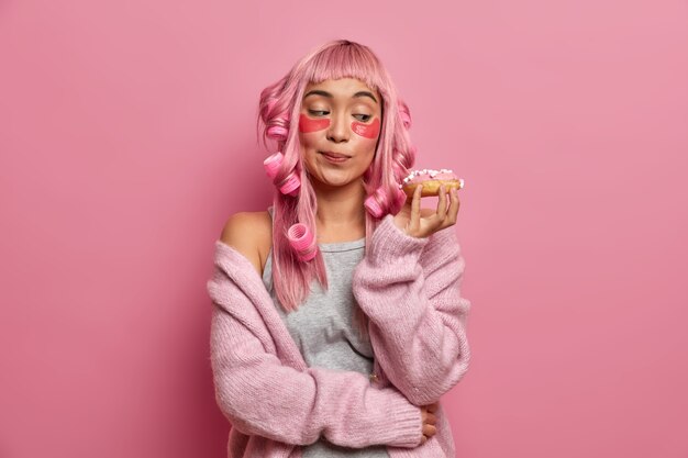 Foto der jungen asiatischen Frau schaut auf appetitlichen Donut, macht Haare mit Rollen, trägt Kollagenpads auf, gekleidet in warmen Pullover