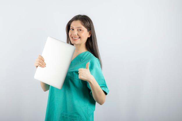 Foto der jungen Krankenschwester mit dem Laptop, der Daumen auf Weiß aufgibt.