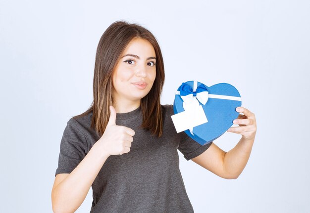 Foto der jungen lächelnden Frau, die sich einen Daumen zeigt und eine herzförmige Geschenkbox hält.