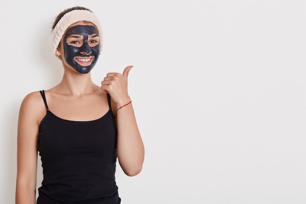 Foto der lächelnden Frau trägt schwarzes T-Shirt und Haarband mit Gesichtsmaske, hat Schönheitsverfahren zu Hause, positiven Ausdruck, zeigt beiseite mit Daumen lokalisiert über weißer Wand.