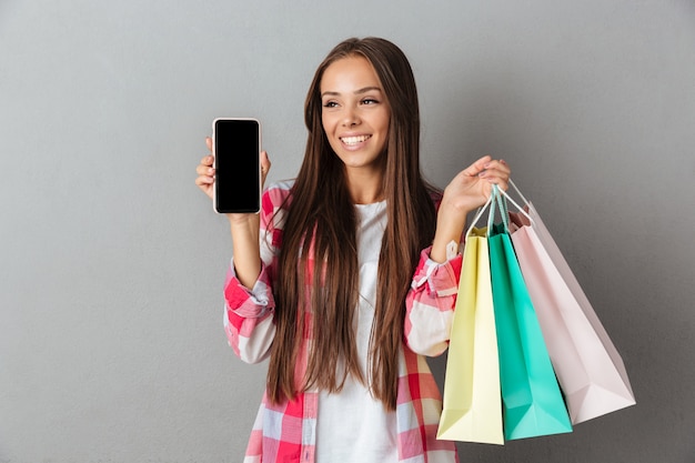 Foto der lächelnden jungen Brünette, die Einkaufstaschen hält, den leeren mobilen Bildschirm zeigt und wegschaut