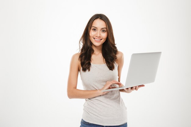Foto der netten Frau mit dem langen braunen Haar, welches den silbernen Personal-Computer aufwirft auf der Kamera, lokalisiert über weißer Wand hält