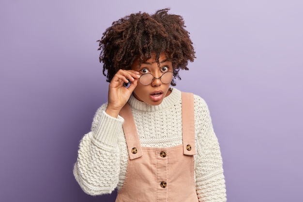 Foto der neugierigen dunkelhäutigen Dame sieht überraschend neugierig durch runde Brille, kann nicht an schockierende Nachrichten glauben, trägt übergroßen weißen Pullover und Overalls, steht über lila Wand