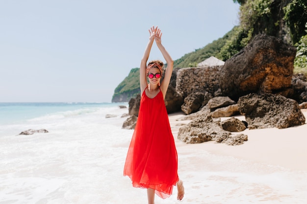 Foto der niedlichen Frau im langen roten Kleid lustiges Tanzen am Sandstrand in voller Länge. Inspiriertes Mädchen in der rosa Sonnenbrille, die herumalbert, während sie sich im exotischen Resort ausruht.