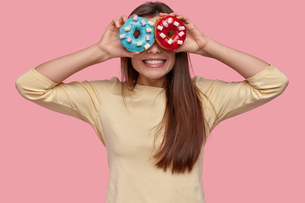 Foto der positiven dunkelhaarigen Frau bedeckt Augen mit zwei Donuts, hat zahniges Lächeln, lässig gekleidet