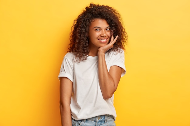 Foto der positiven dunkelhäutigen Frau mit dem knackigen Haar, hat sanftes Lächeln, berührt Kinn, gekleidet in lässigem weißem T-Shirt und Jeans, steht gegen gelbe Wand.