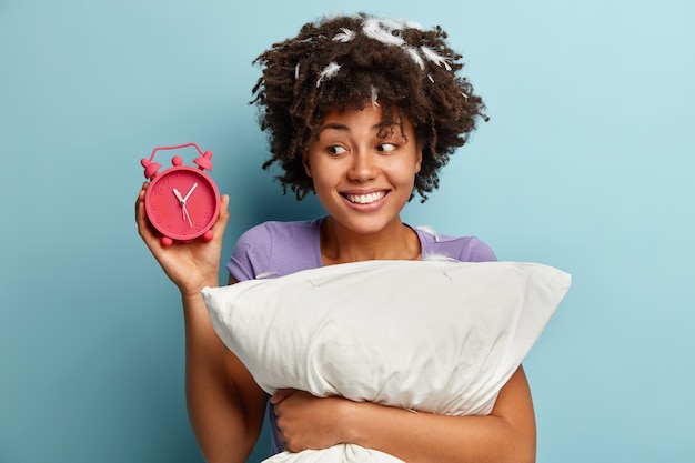 Foto der reizenden afroamerikanischen Frau mit dem lockigen Haar, zeigt Zeit auf Wecker, froh, langen Schlaf nachts zu haben, lächelt positiv, hält weiches weißes Kissen, isoliert über blauer Wand. Schlafen