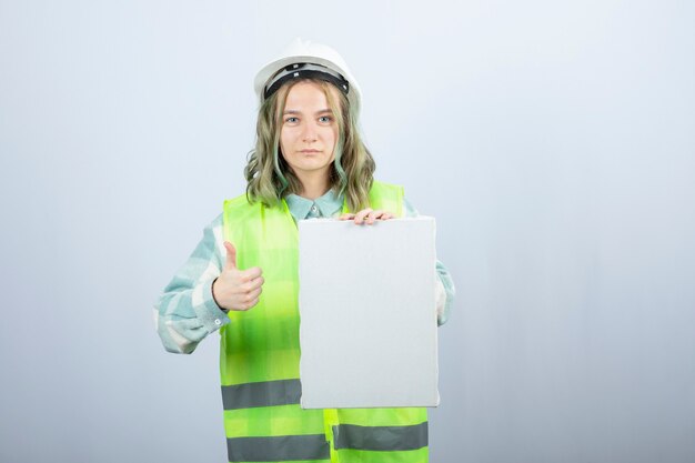 Foto der schönen Ingenieurin, die leere Leinwand hält und Daumen aufgibt. Hochwertiges Foto