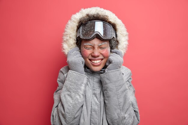 Foto der überglücklichen Frau trägt Kapuze der grauen Jacke lächelt angenehm hat rotes Gesicht mit Frost bedeckt geht Skifahren im Dezember.