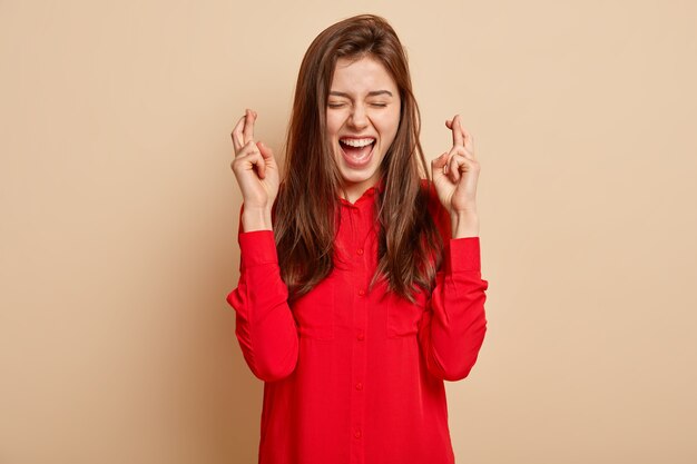 Foto der überglücklichen schönen dunkelhaarigen Frau kreuzt die Finger zum Glück, glaubt aufrichtig an den Sieg, hält die Augen geschlossen, in rote Bluse gekleidet, Gesten über beige Wand. Körpersprache