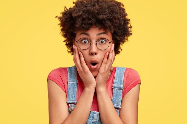 Foto der überraschten afroamerikanischen Frau berührt Wangen, öffnet Augen und Mund weit, gekleidet in Freizeitkleidung, isoliert über gelber Wand. Schockierte Mischlingsfrau posiert allein drinnen.