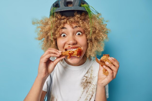 Foto der überraschten lockigen jungen Frau, die ungesundes Essen isst, hält gebratene Nuggets mit Ketchup, die sehr hungrig sind, trägt einen Schutzhelm und ein weißes, schmutziges T-Shirt, das über blauem Hintergrund isoliert ist