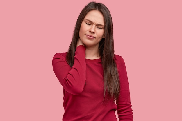 Foto der unzufriedenen Frau hält Hand am Hals, Augen geschlossen, hat lange Haare, trägt roten Pullover
