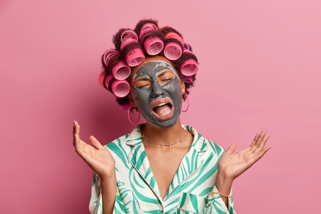 Foto der unzufriedenen weinenden hausfrau fühlt sich müde und erschöpft, trägt lockenwickler und schönheitsgesichtsmaske, spreizt handflächen, gekleidet in freizeitkleidung, isoliert auf rosa. kosmetologie, spa-behandlung