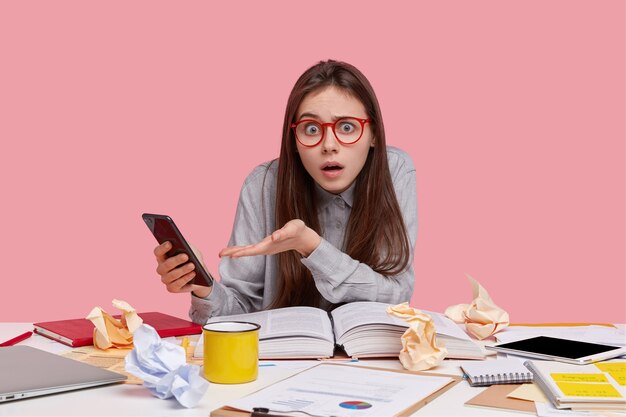 Foto der verwirrten emotionalen Dame trägt eine Brille, hält modernes Handy, fühlt sich verwirrt, liest wissenschaftliche Literatur, verblüfft, um Benachrichtigung zu erhalten